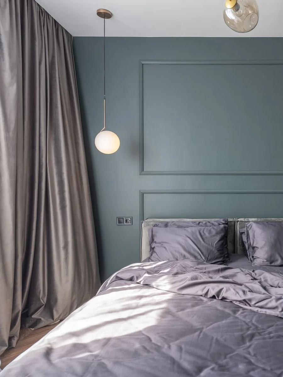 White air purifier on a bedroom floor beside a bed with linen bedding and a floor lamp casting soft light