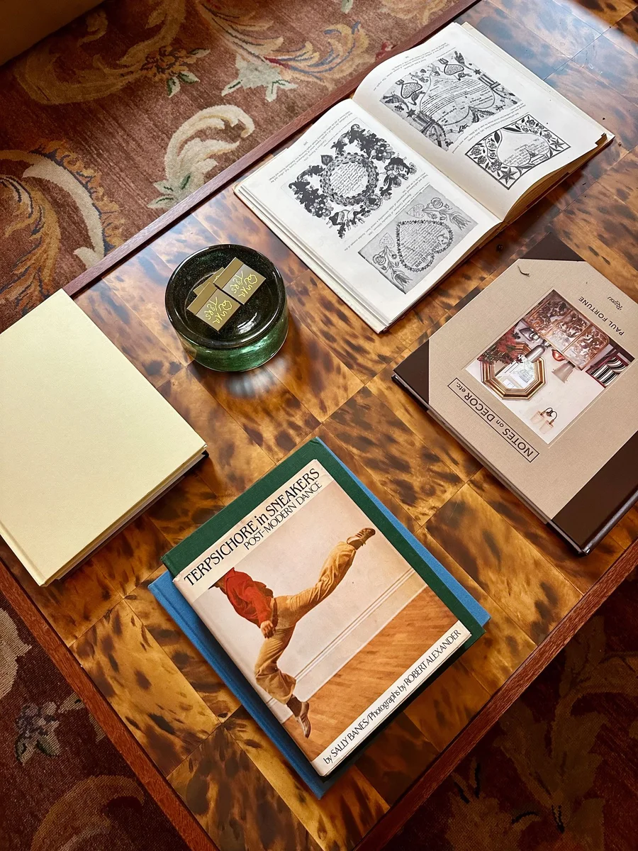 Stack of oversized hardcover books and a round wood tray holding two cups on a walnut coffee table in a bright living room
